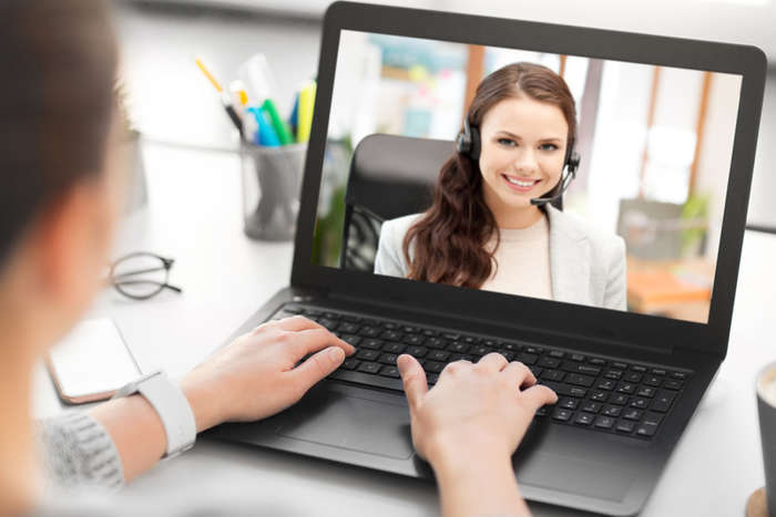 Betrachter sieht von hinten an Frau vor Laptop vorbei auf den Bildschirm. Dieser zeigt eine Frau, die ihn anschaut und lächelt.