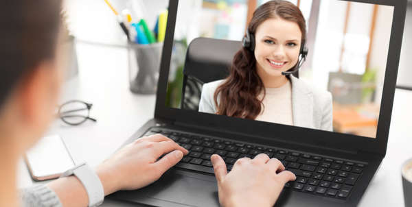 Betrachter sieht von hinten an Frau vor Laptop vorbei auf den Bildschirm. Dieser zeigt eine Frau, die ihn anschaut und lächelt.